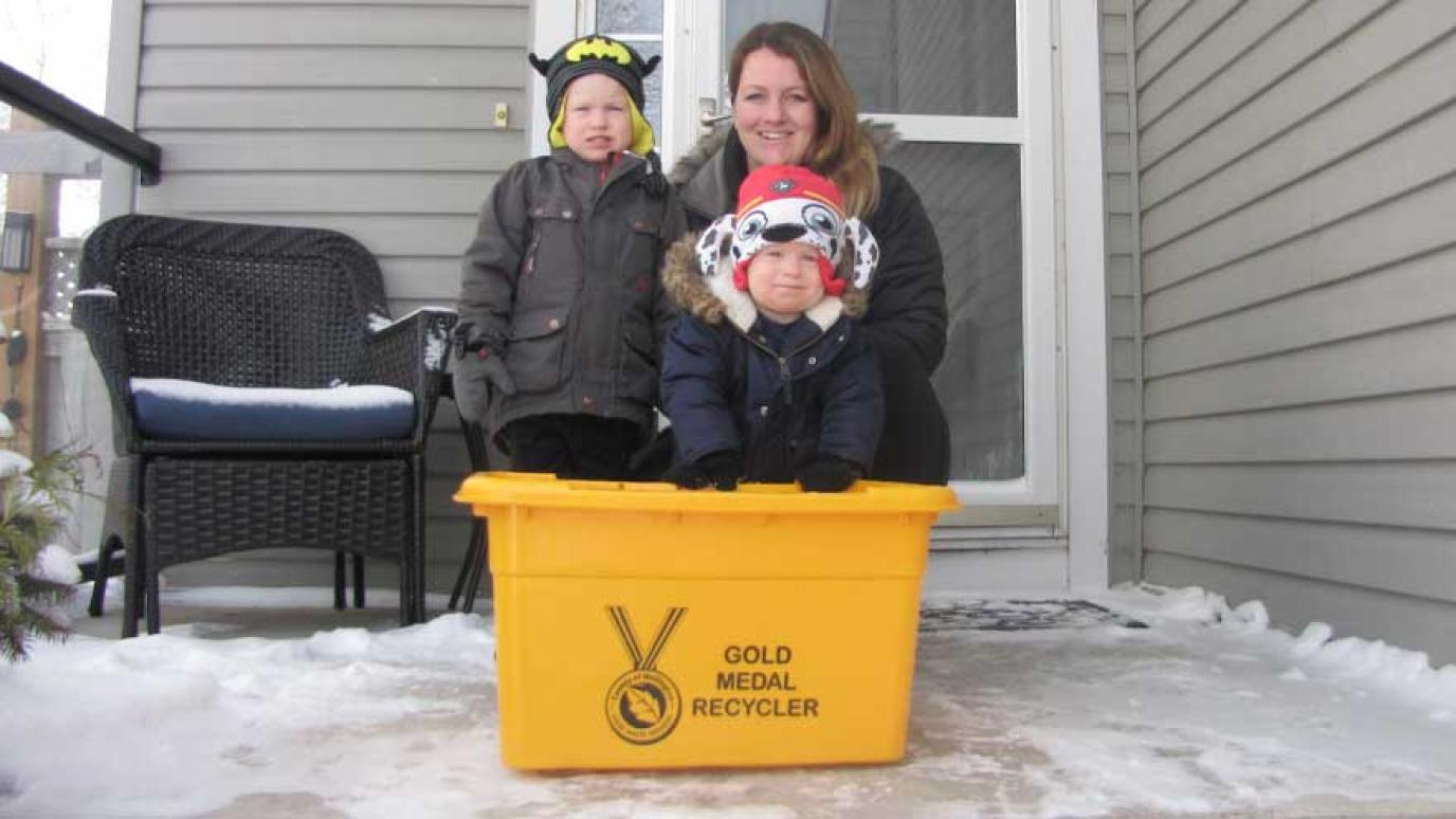 Woman and two children posing happily on porch next to new gold recycling bin
