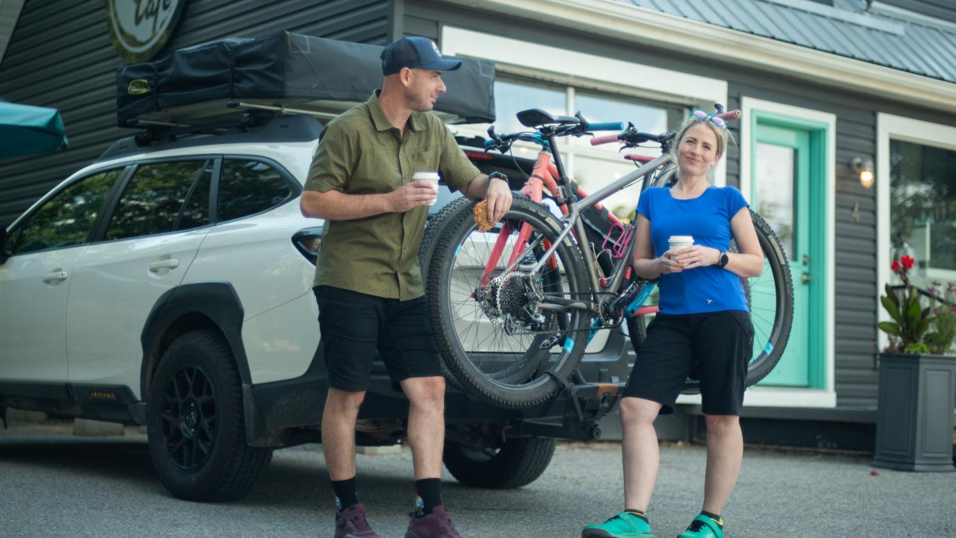 two people outside cafe holding coffee leaning against truck with bikes on the back