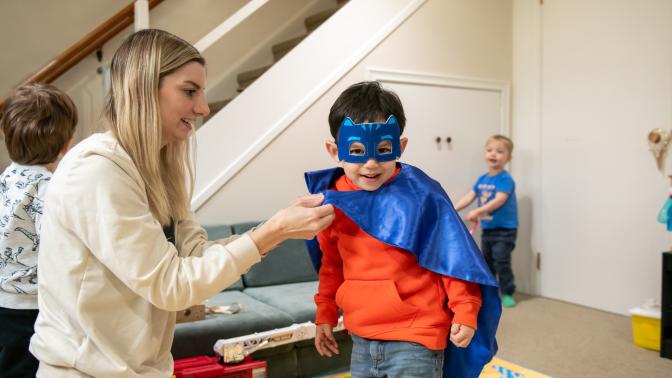 A home childcare provider ties a cape onto a young boy wearing super hero mask at her home child care. 