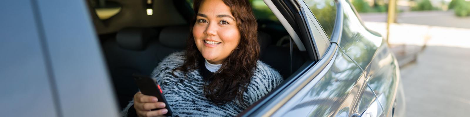 Smiling young woman holding a smartphone peering out the window from backseat of rideshare vehicle